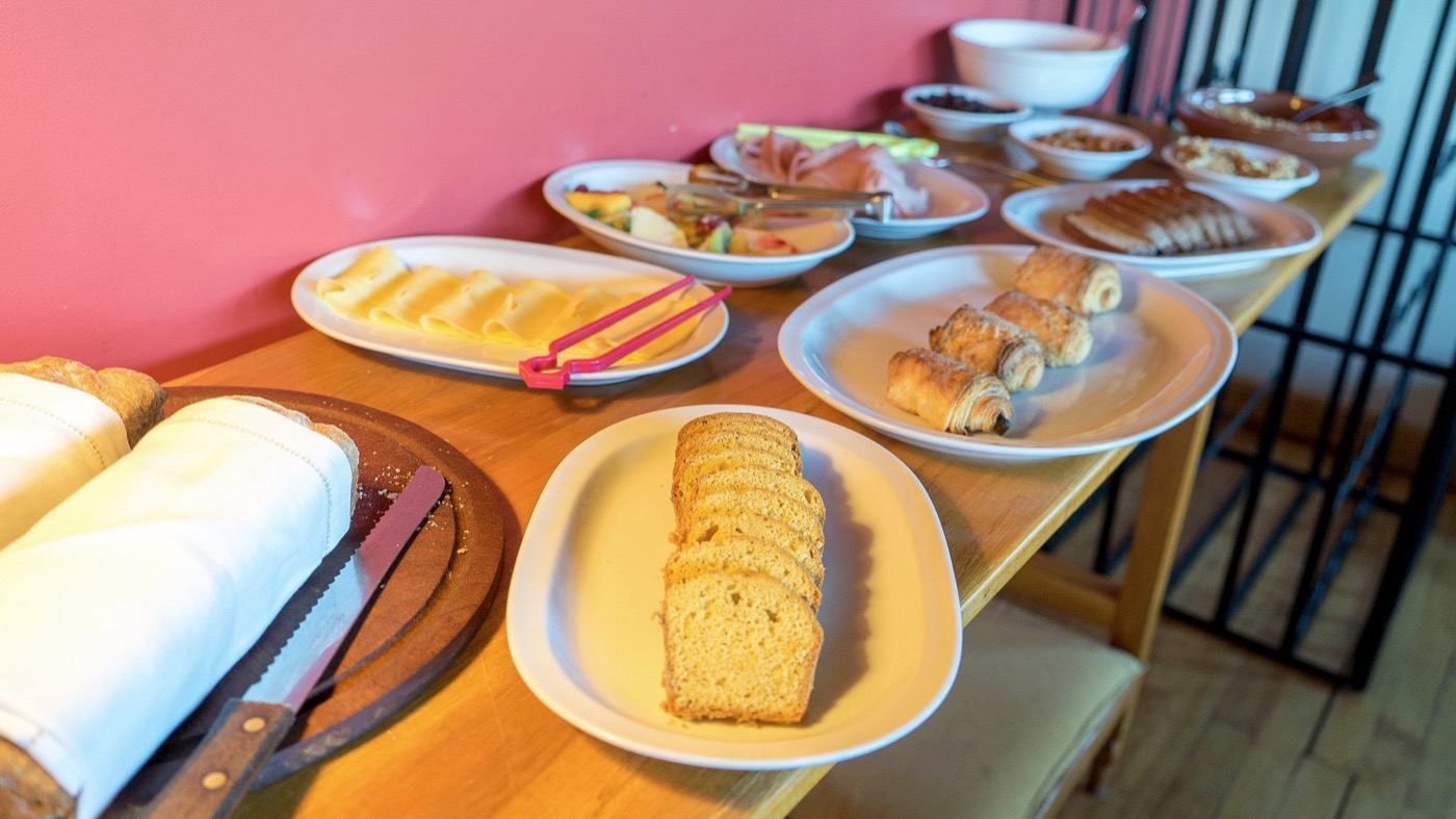 A continental breakfast spread on a wooden table: sliced bread, cheese, fruit, ham, pastries, and cereals
