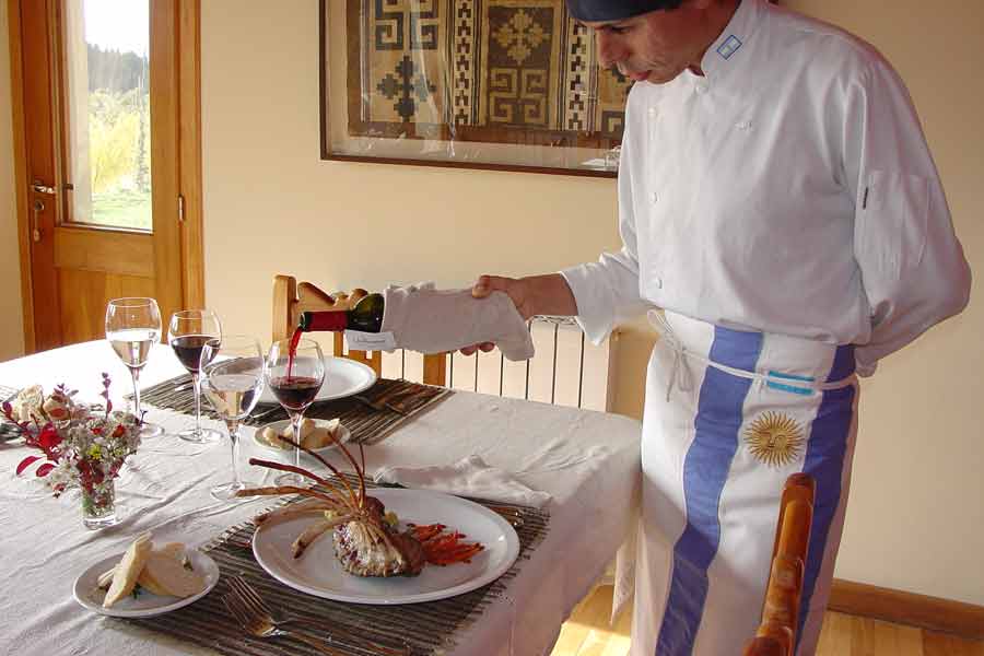A chef in whites and an Argentine-flag apron pouring red wine at a set table beside a plated dish in the lodge dining room