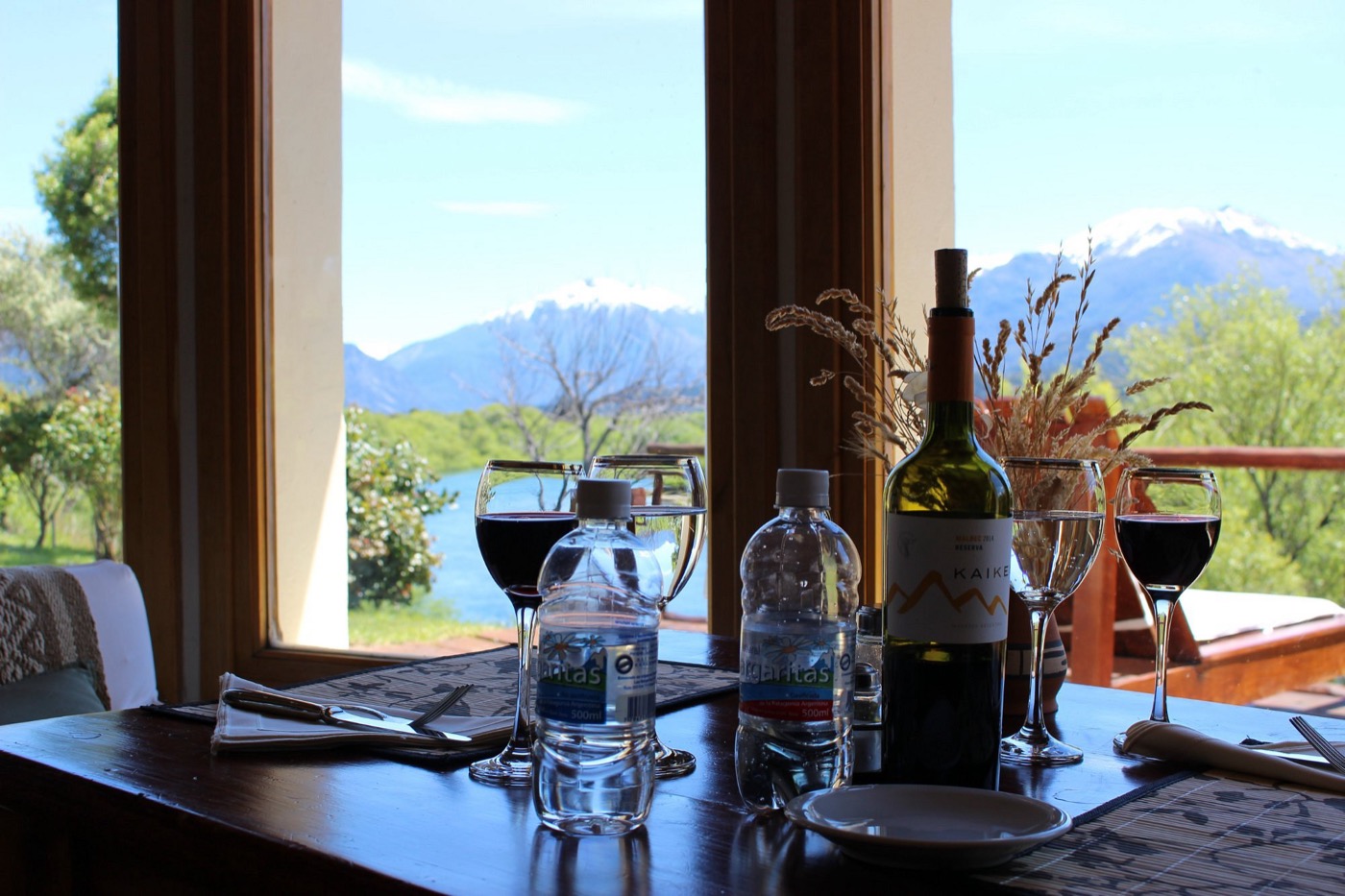 A dining table set with red wine, water bottles, and dried grasses in front of tall windows looking out to a river and snow-capped mountains