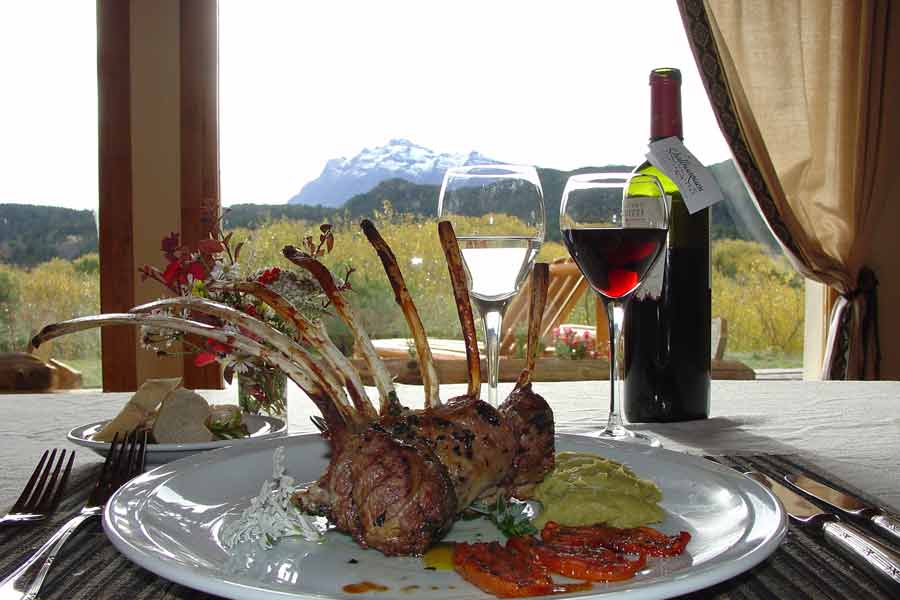 A plated rack of lamb with mashed potatoes and roasted tomatoes beside glasses of red and white wine, with a snow-capped mountain through the window