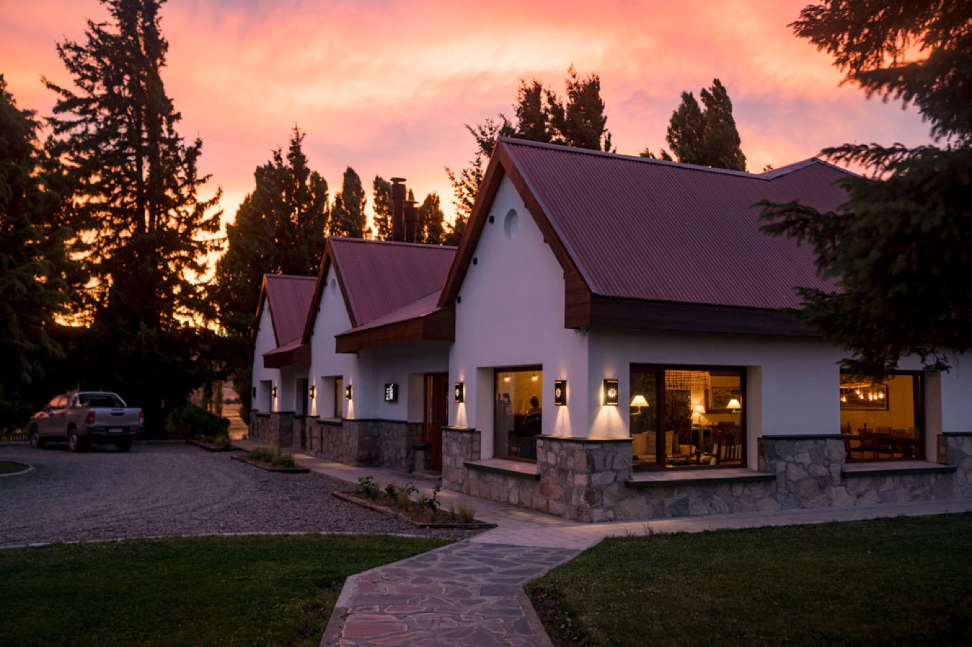 White lodge buildings with dark red gabled roofs glowing at sunset, a pickup truck parked on the gravel drive