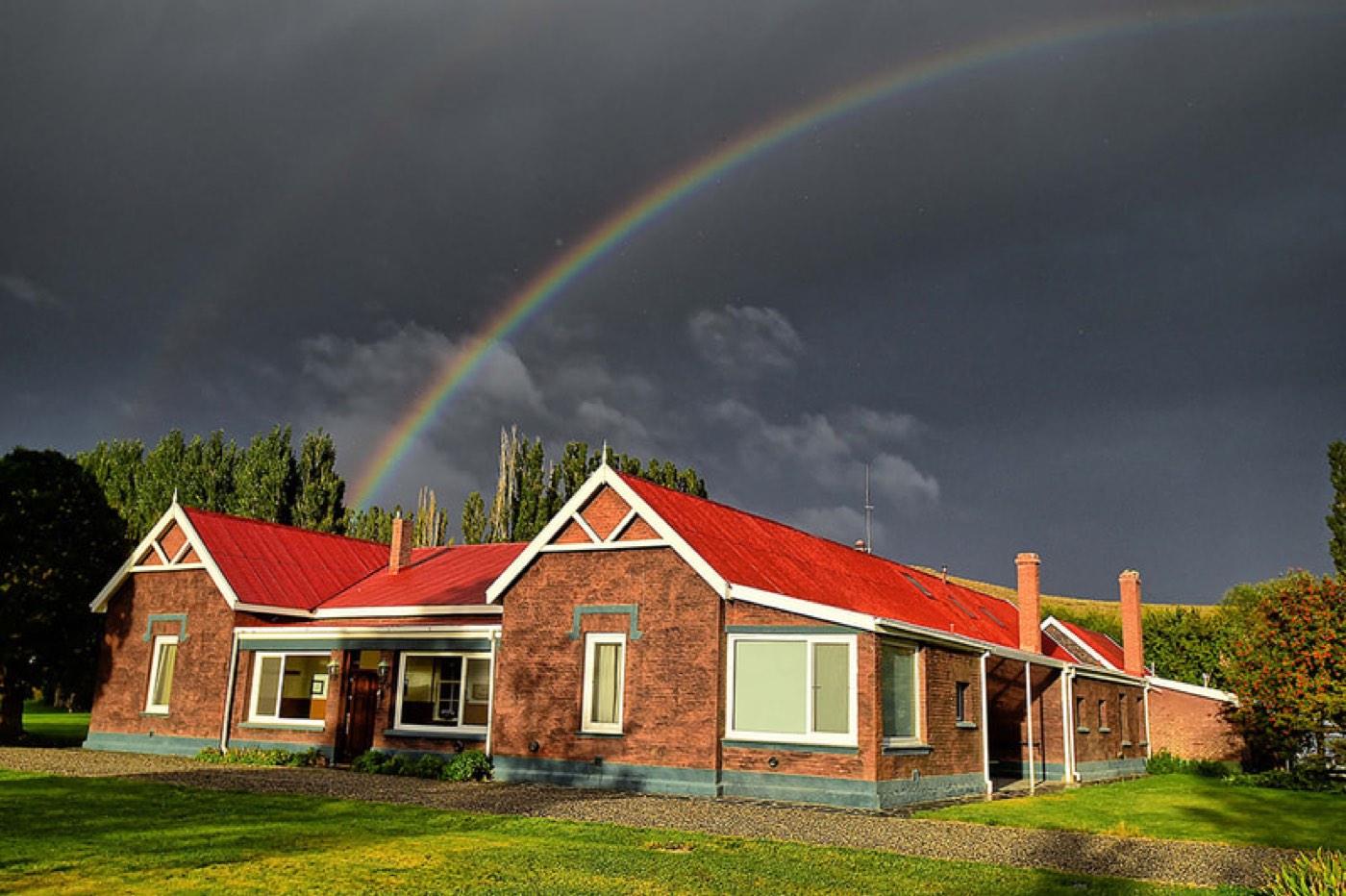 A red-roofed brick ranch building beneath a vivid double rainbow arcing across a stormy sky