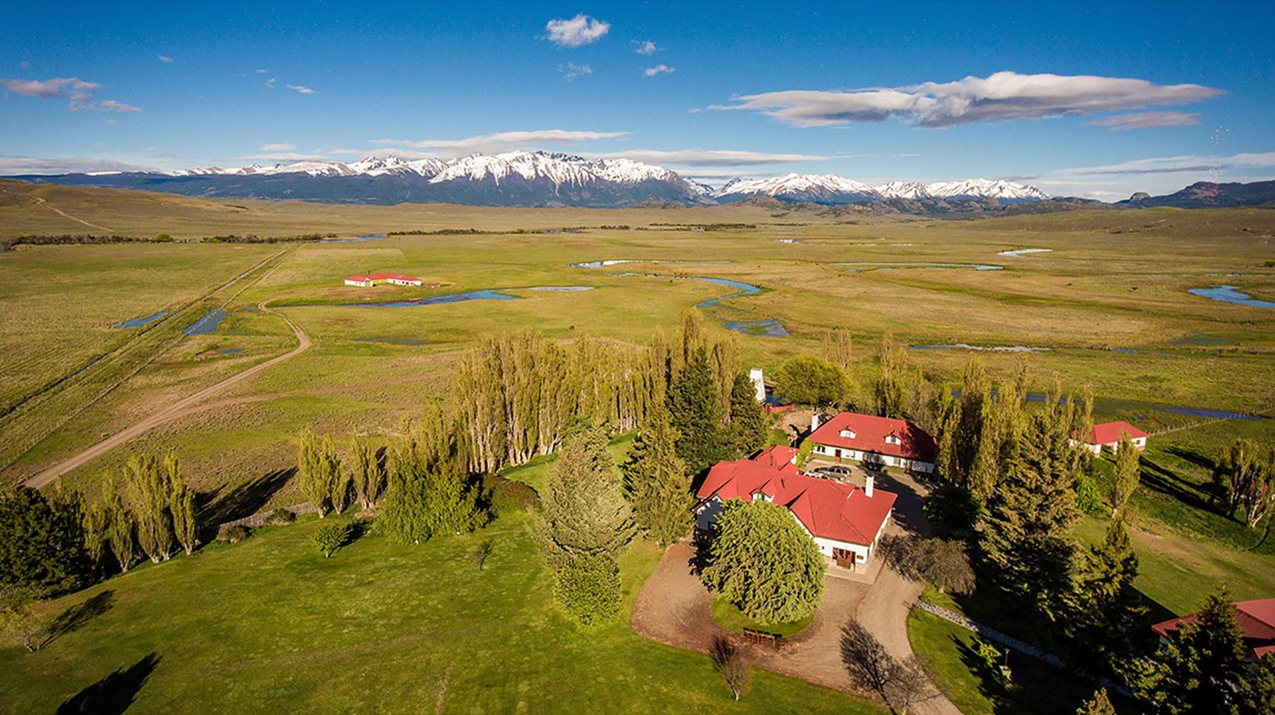 An aerial view of the lodge's red-roofed buildings amid green lawns, with wetlands stretching to snow-capped mountains