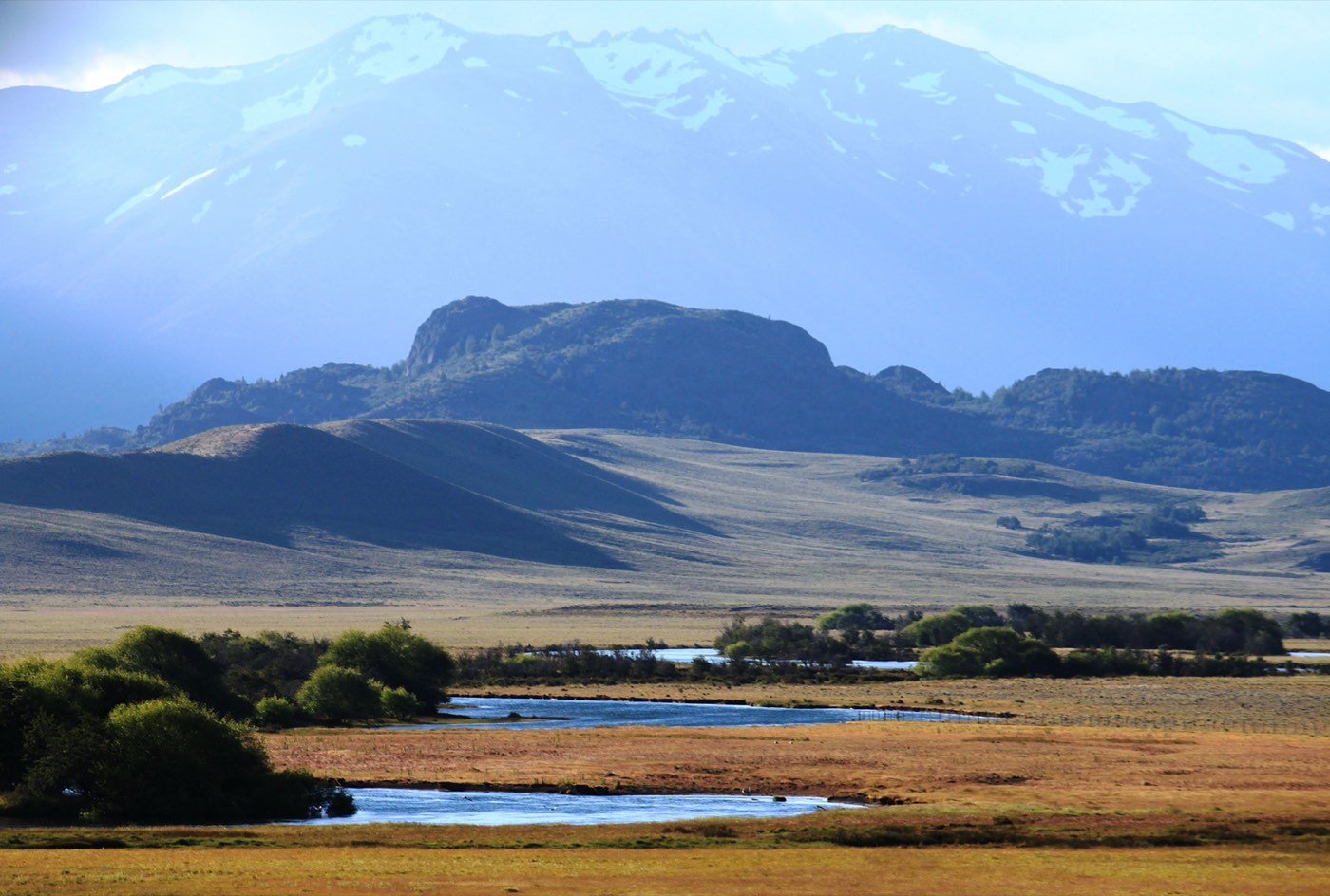 A hazy, backlit mountain range with a rounded rock outcrop rising over golden grassland and a river