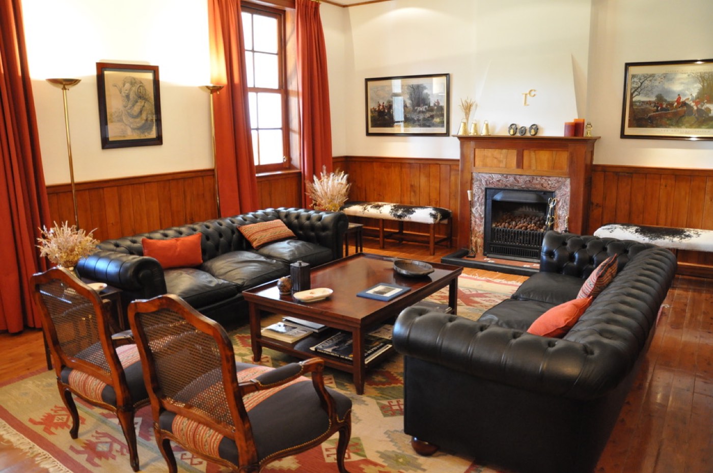 A formal lounge with black leather chesterfield sofas, equestrian prints, and a fireplace beneath a brass monogram