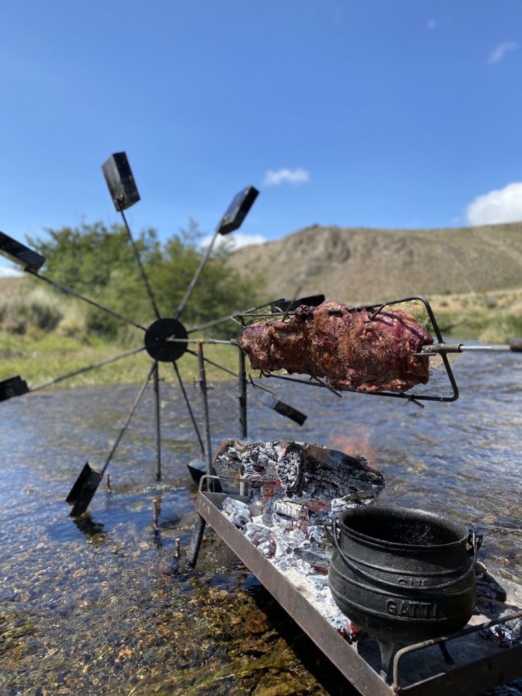 A cut of meat on a spit roasting over an open fire built in the shallows of a river