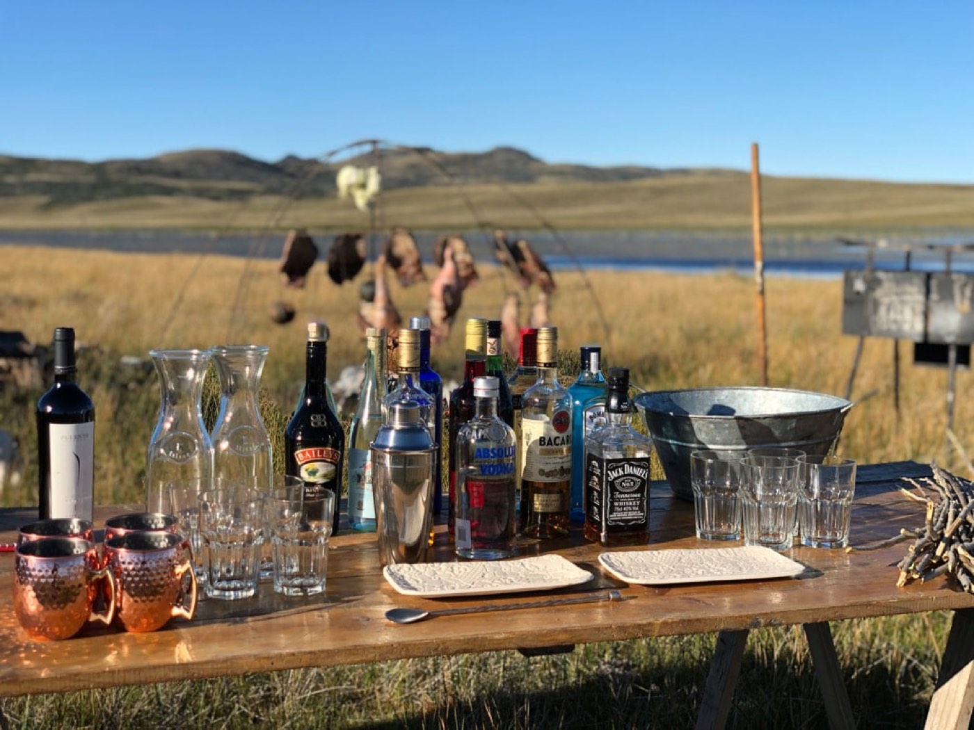 An outdoor bar set with wine, spirits, and copper mugs on the Patagonian steppe, dried game hanging to cure behind it