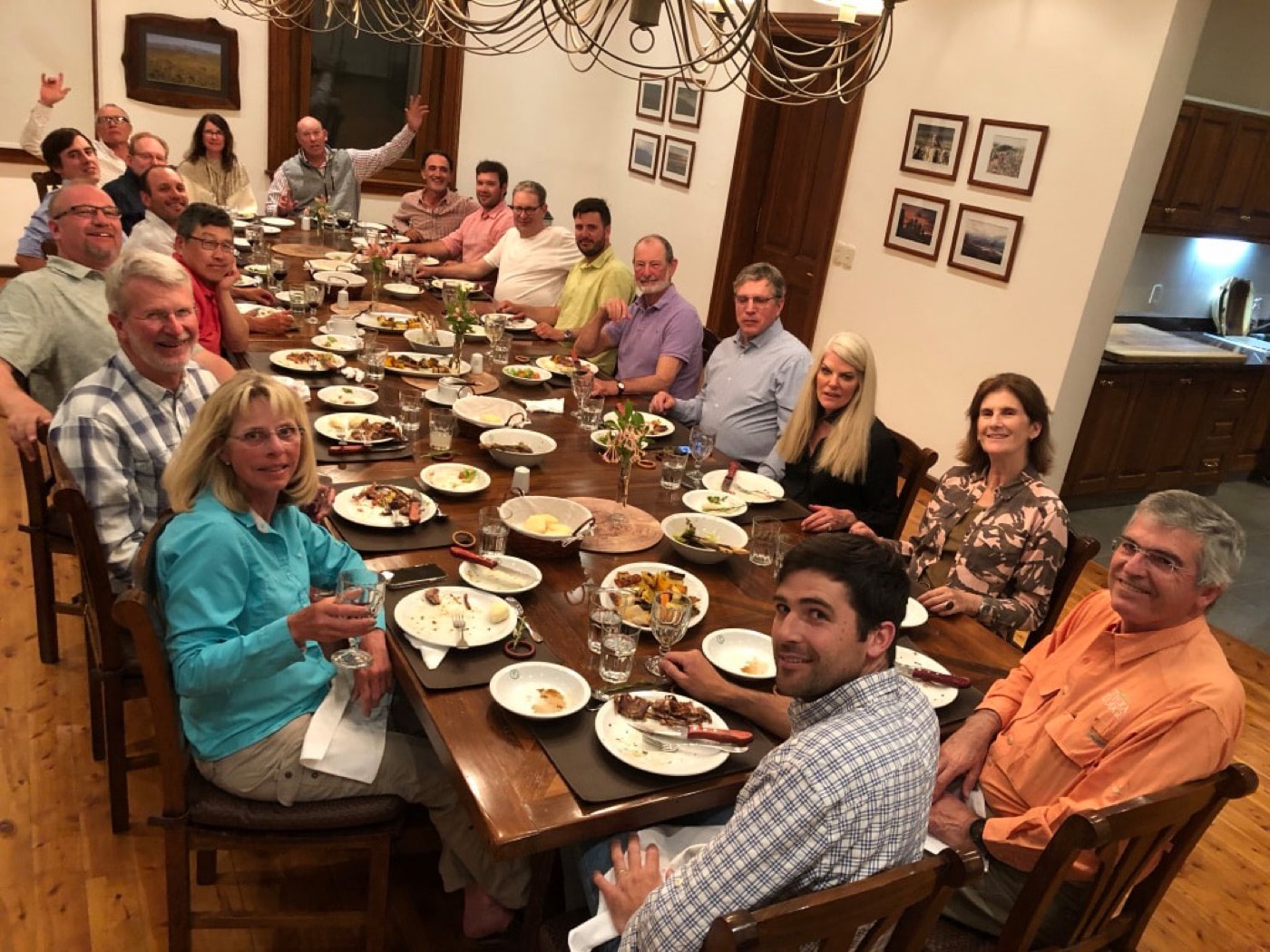 A large group of guests seated around the lodge's long dining table, mid-dinner, under a wrought-iron chandelier