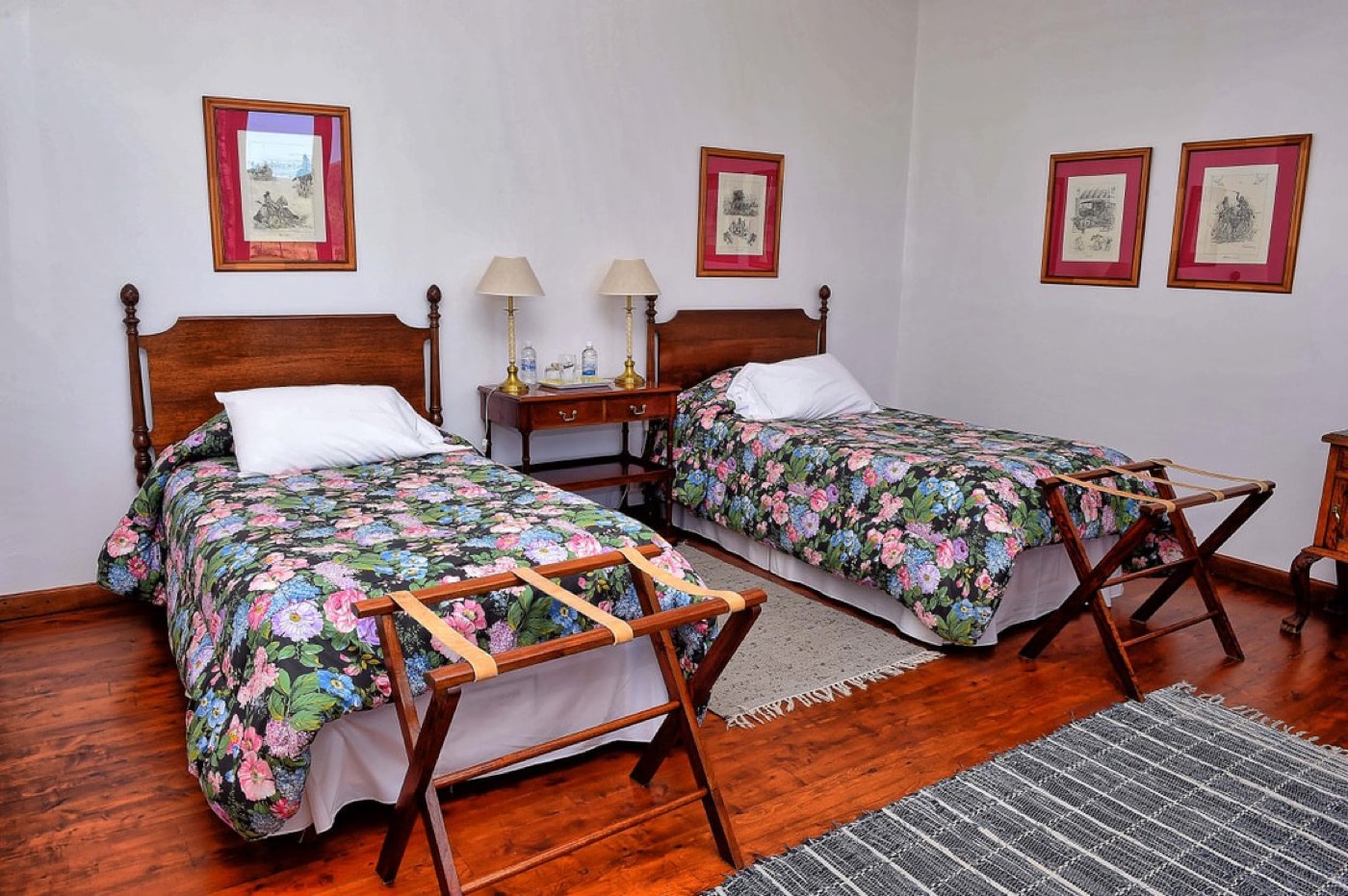 A twin room with pink floral bedspreads, vintage equestrian prints, and wooden luggage racks
