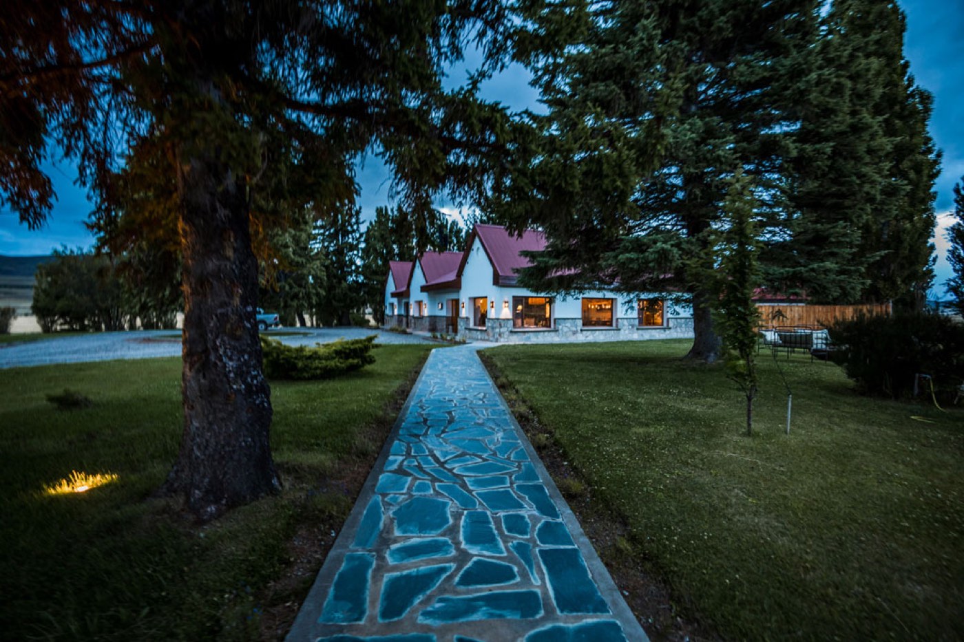 A blue-tiled stone walkway leading to the lit lodge at dusk, conifers standing on either side
