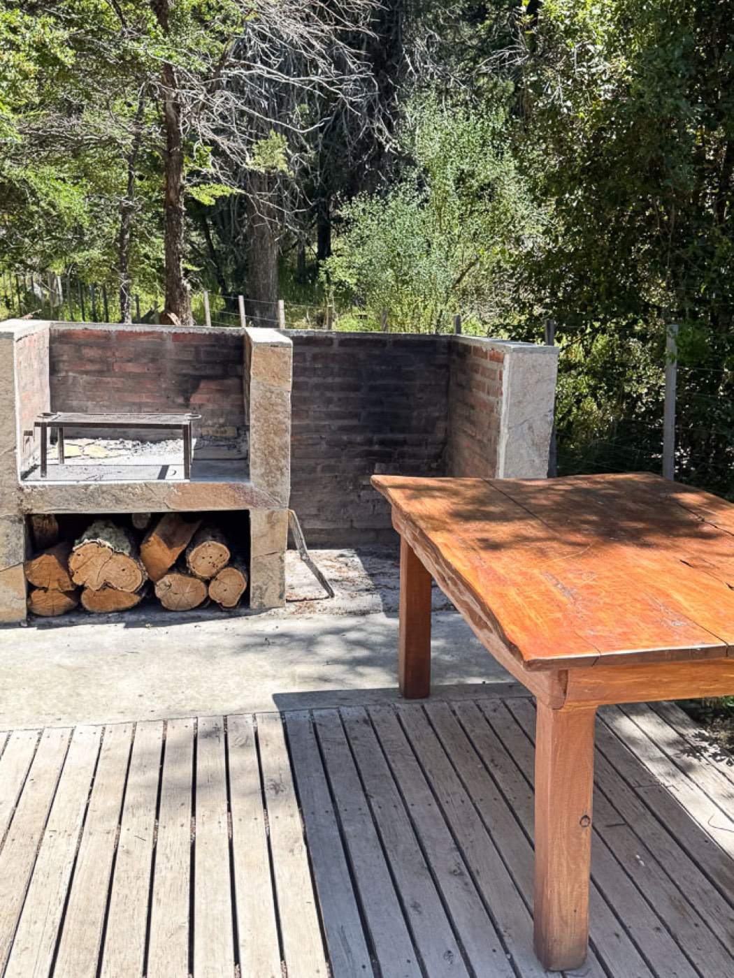 An outdoor stone barbecue pit stacked with firewood, beside a rustic wooden table on a shaded deck