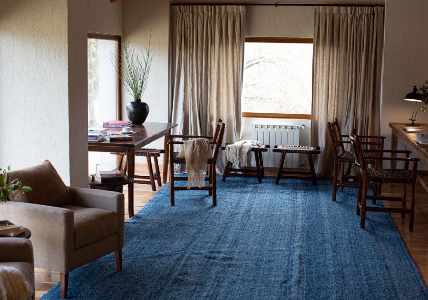 A living-room reading nook with a blue woven rug, an upholstered armchair, and a writing desk stacked with books