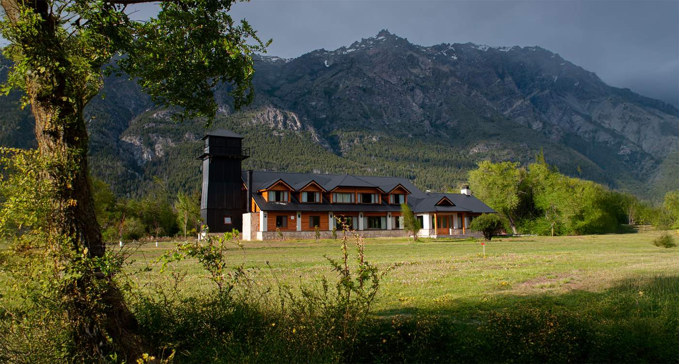 The lodge's two-story timber-and-stone main building and dark lookout tower, set on a green meadow beneath a steep, snow-dusted Andean ridge