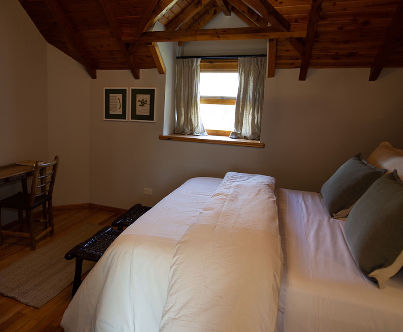 A cozy attic bedroom with a peaked wood-beam ceiling, a writing desk, and a window overlooking the grounds