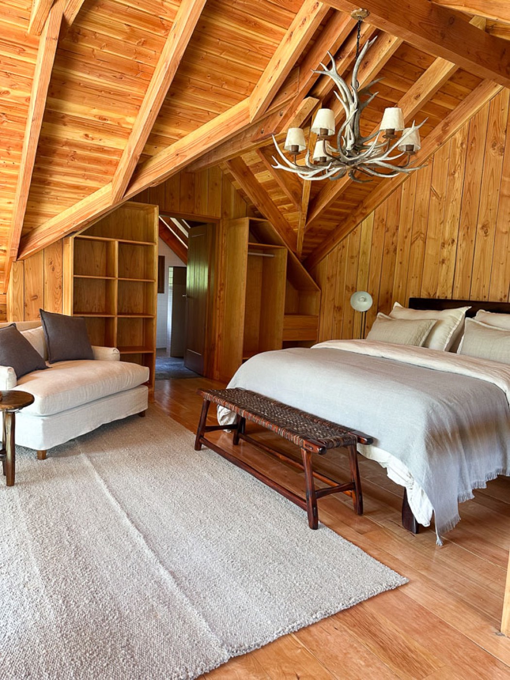 A loft bedroom beneath a steep timber roofline, with an antler chandelier and built-in shelving