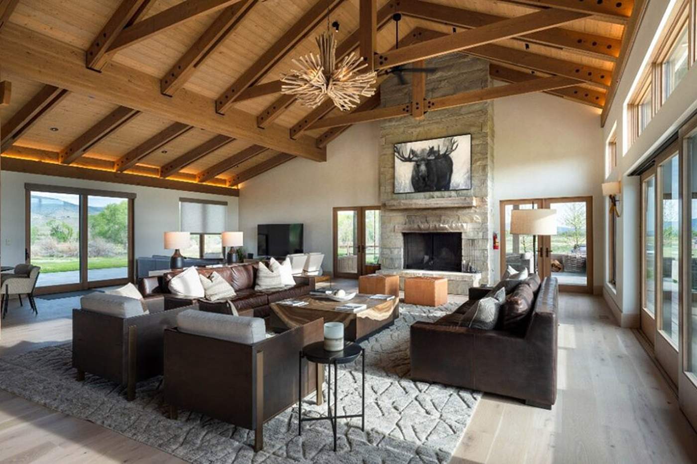 A great room with a vaulted timber-beam ceiling, a tall stone fireplace, and brown leather sofas and armchairs facing floor-to-ceiling windows
