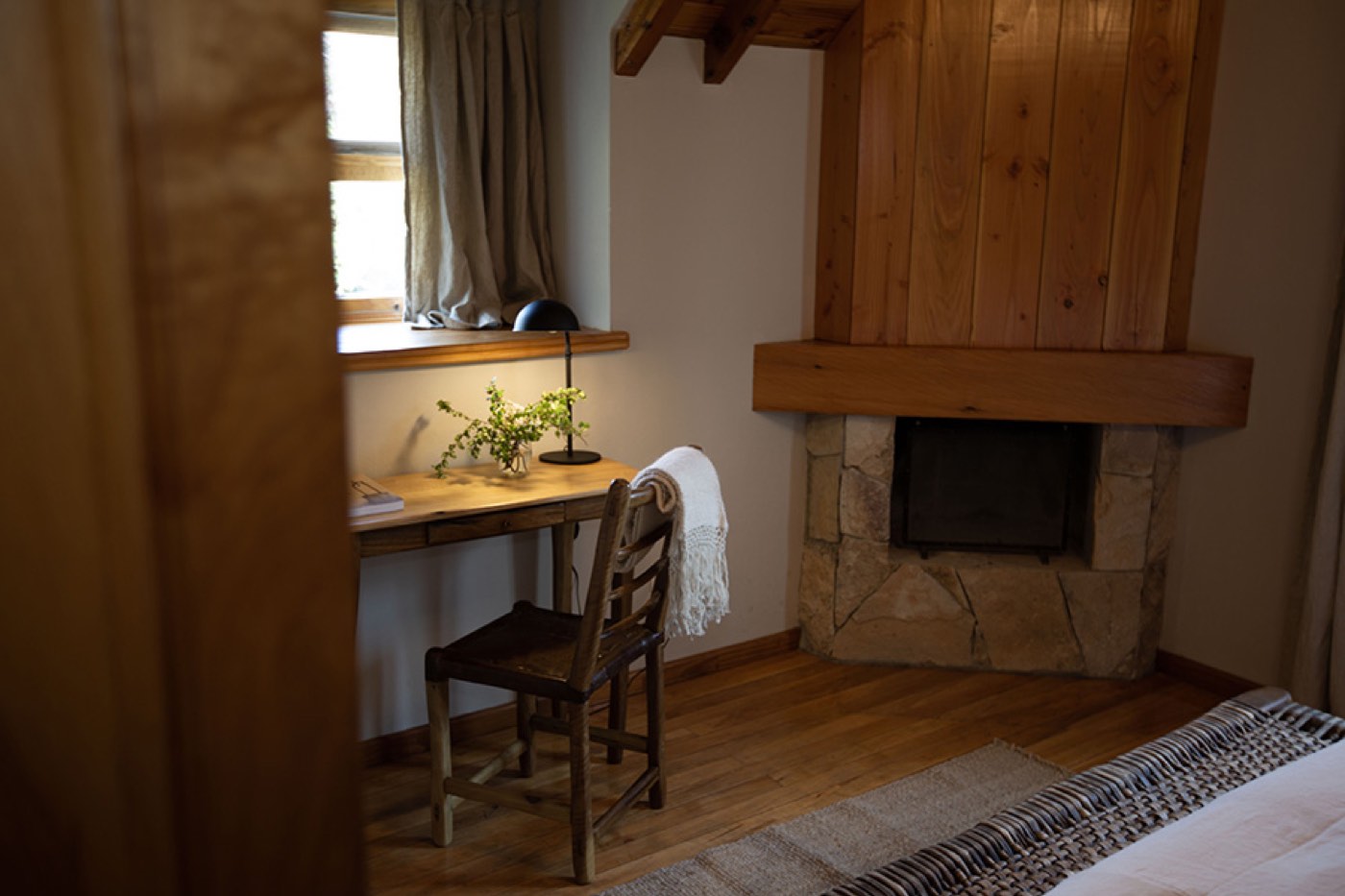 A guest room corner with a small writing desk, a potted plant, and a stone-and-timber fireplace
