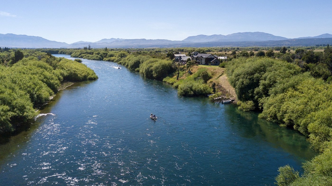 An aerial view of a wide river bending past a lodge on its bank, a small fishing boat drifting mid-river, mountains in the distance