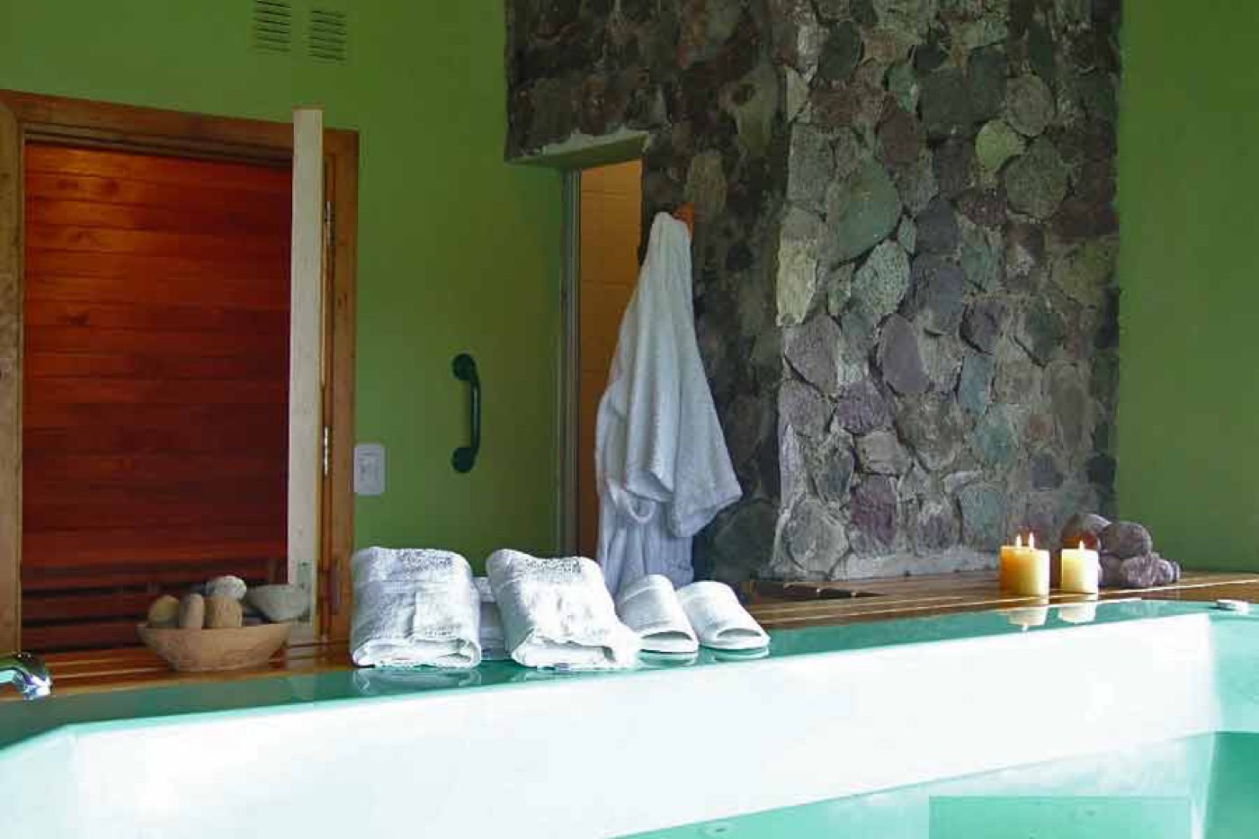 A spa area with a stone accent wall, a hot tub's edge lined with towels and slippers, and a robe hanging by the sauna door