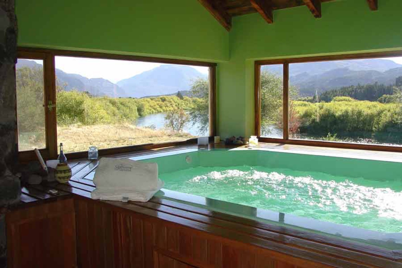 An indoor hot tub with a view of the river and mountains through the windows, a rolled towel and toiletries on its ledge