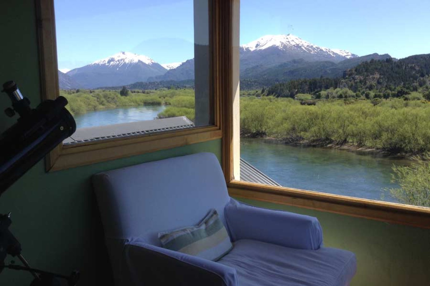 A telescope beside a white armchair, floor-to-ceiling windows framing a river and snow-capped mountains