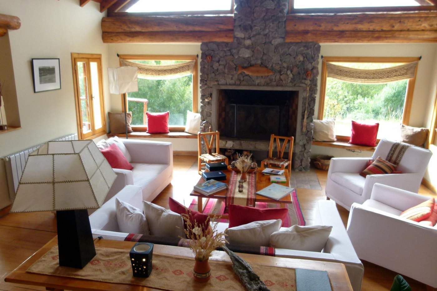 A sunlit great room with skylights and a vaulted ceiling, white sofas with red pillows facing a stone fireplace topped with a carved wooden fish
