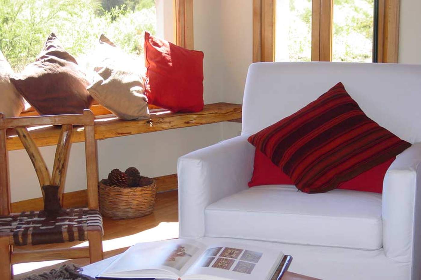 A window-seat corner with woven and striped pillows, beside a white armchair and a basket of pinecones