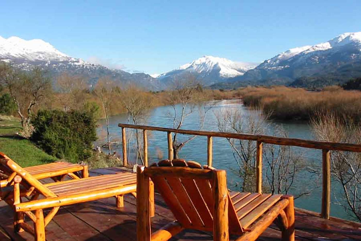 A wooden deck with log-frame lounge chairs overlooking a river, bare autumn trees along the bank and mountains beyond