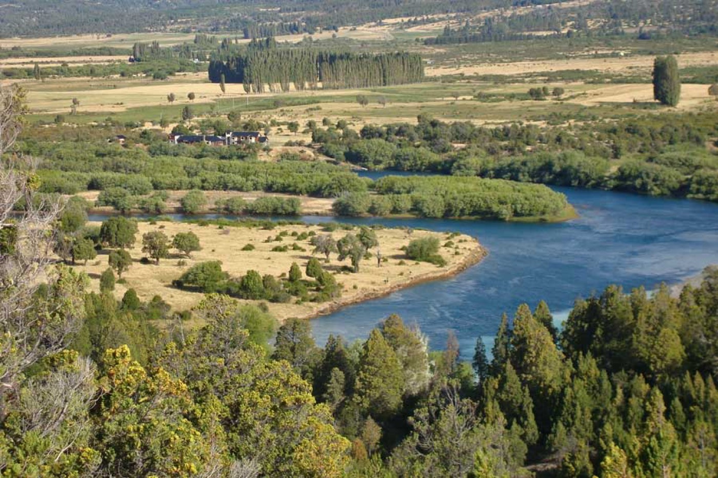 An elevated view of a river curving through a green valley, a lodge among trees on the far bank, poplar windbreaks and farmland beyond