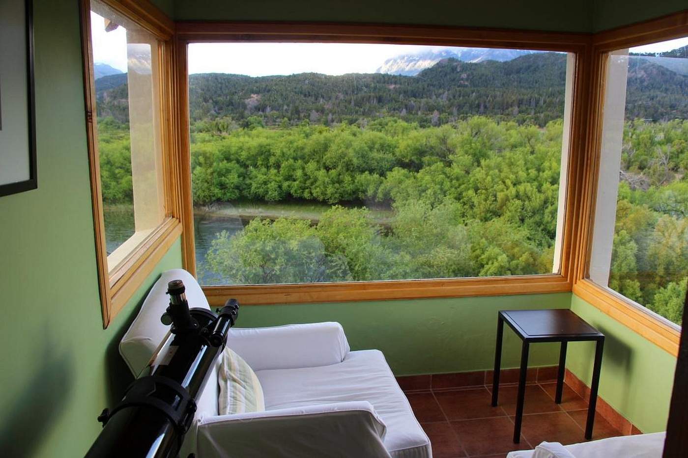 A green-walled sunroom with a telescope, a white daybed, and large windows overlooking the river and forest