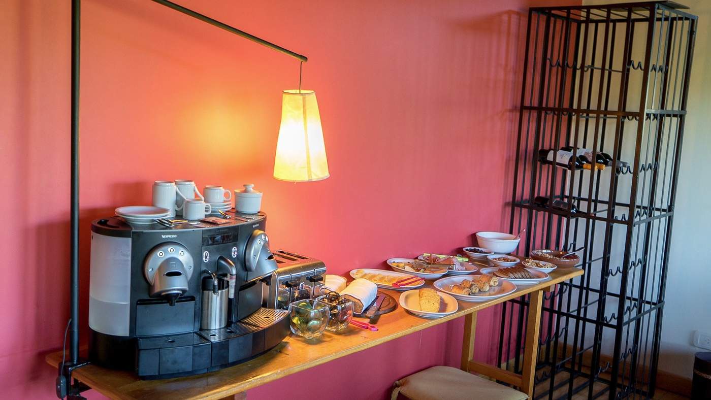 A breakfast station with an espresso machine, a hanging pendant lamp, and a wine rack against a coral-pink wall