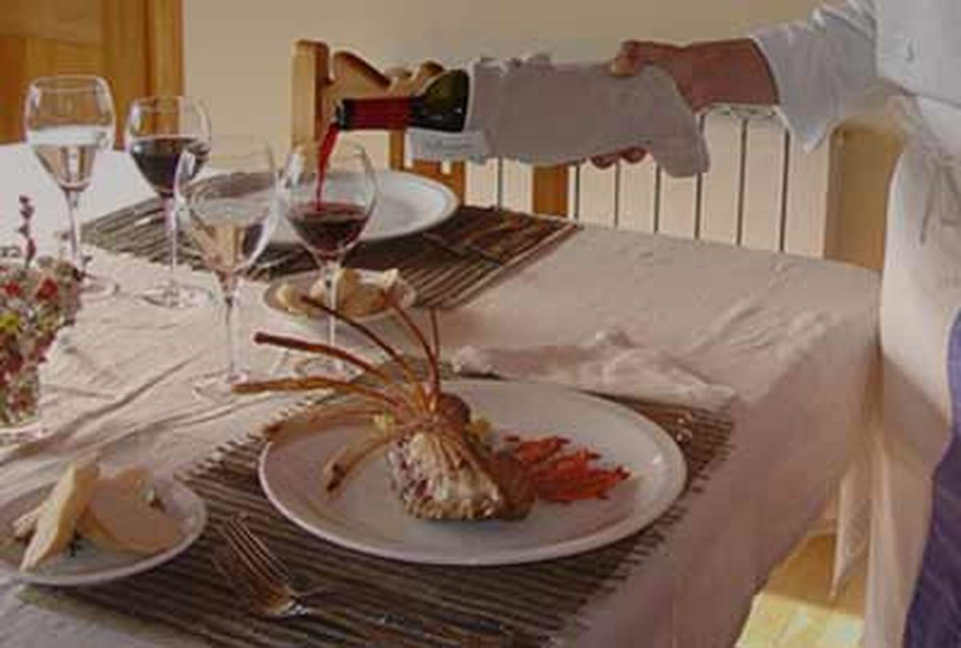 A chef pouring red wine tableside beside a plated rack of lamb, a vase of wildflowers and bread set alongside
