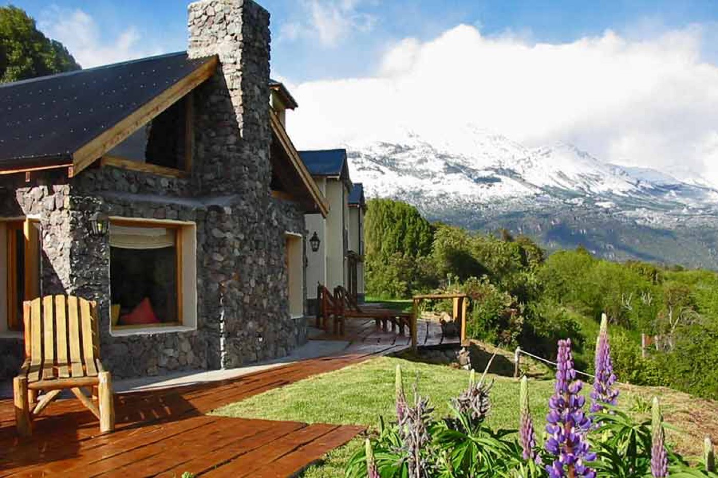 A stone-and-timber lodge exterior with a steep black roof, an outdoor patio, and purple lupines blooming in the foreground, snow-capped mountains behind