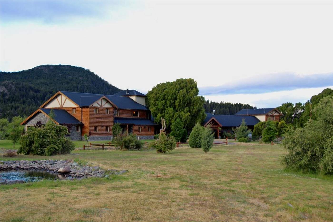 A wide view of the log lodge complex beside a small pond, forested hills behind under evening light