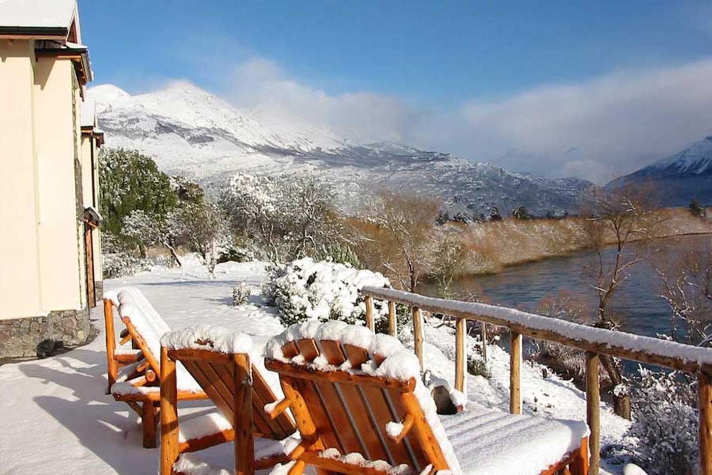 A snow-covered deck with log-frame lounge chairs, overlooking a river beneath snow-dusted mountains