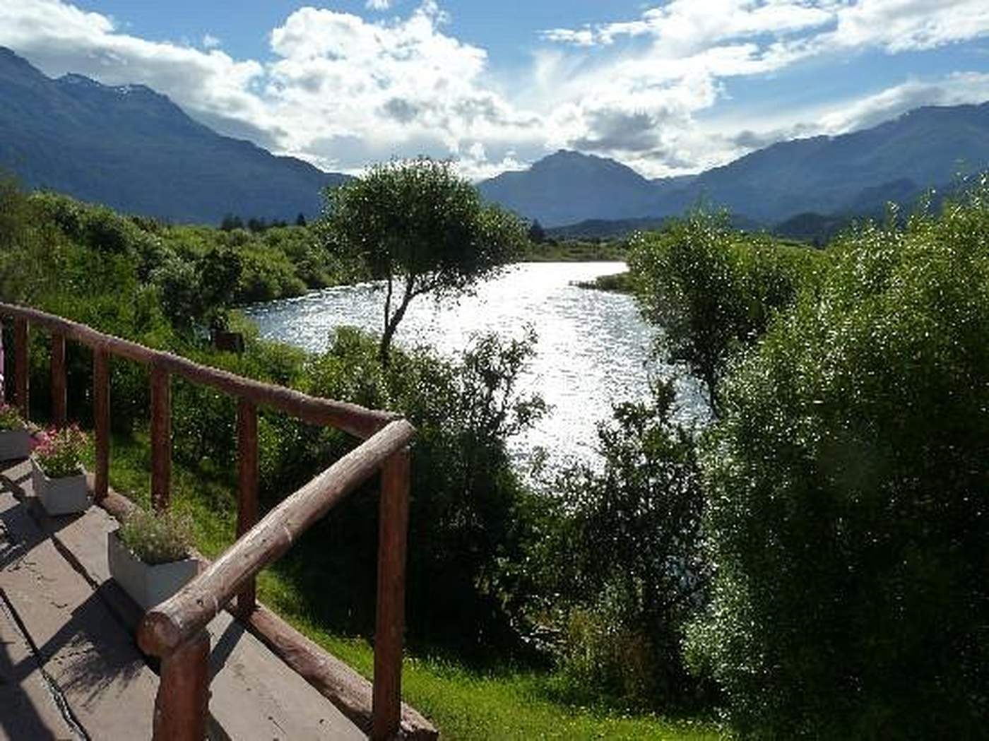 A wooden deck railing with potted flowers, overlooking a sunlit river bend and distant mountains