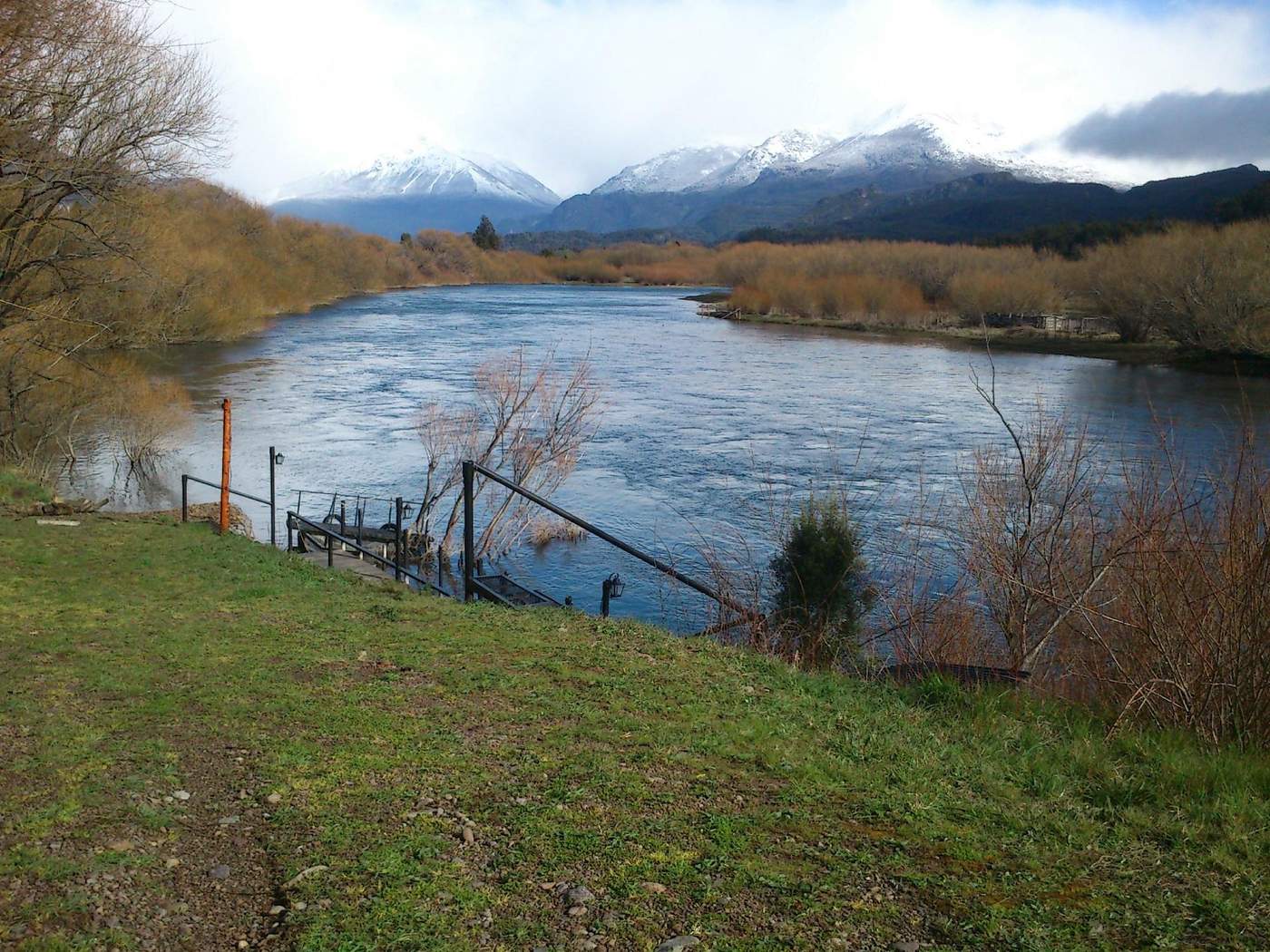 A view of a river bend from the grassy bank, a small dock leading to the water and snow-capped mountains under a cool sky