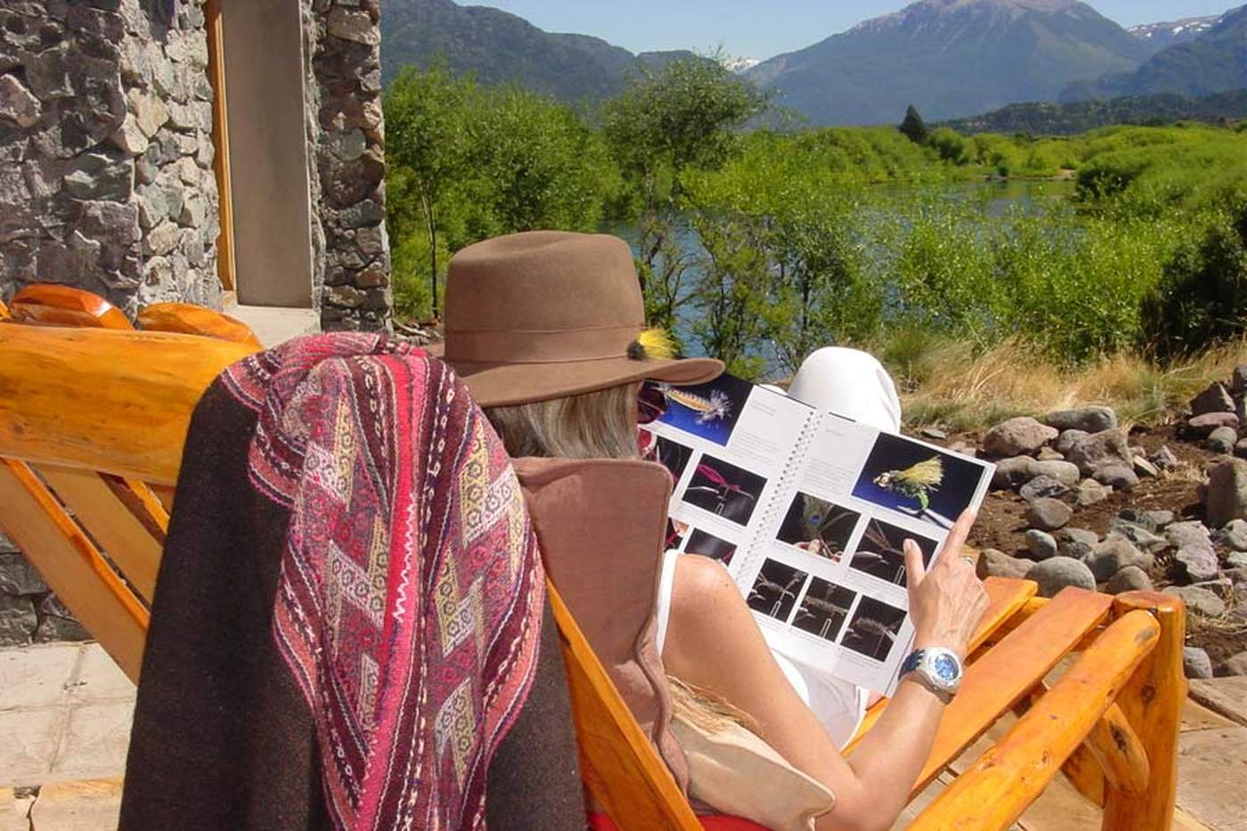 Two guests relaxing in log-frame lounge chairs on the porch, reading a fly-pattern catalog together, a river and mountains beyond