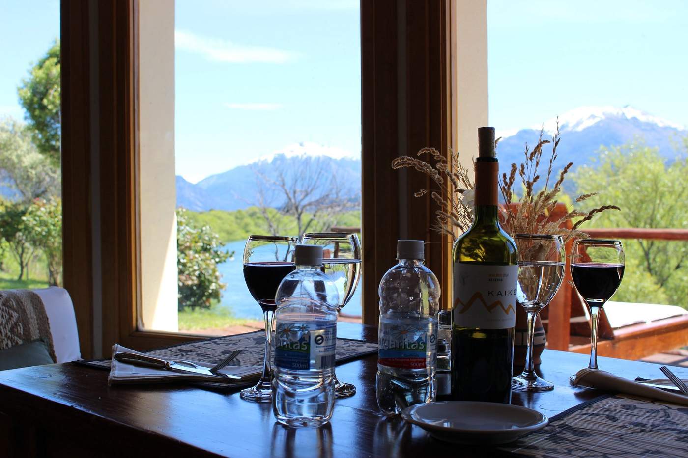 A dinner table set beside a large window, wine and sparkling water poured, a river and snow-capped mountains outside