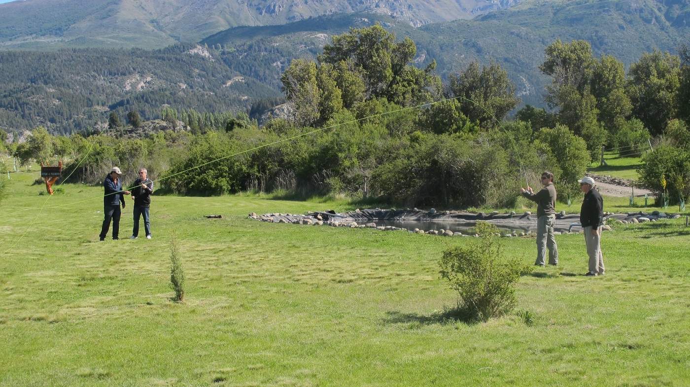 Four anglers practicing their casting on the lawn beside a small pond, mountains and forest behind