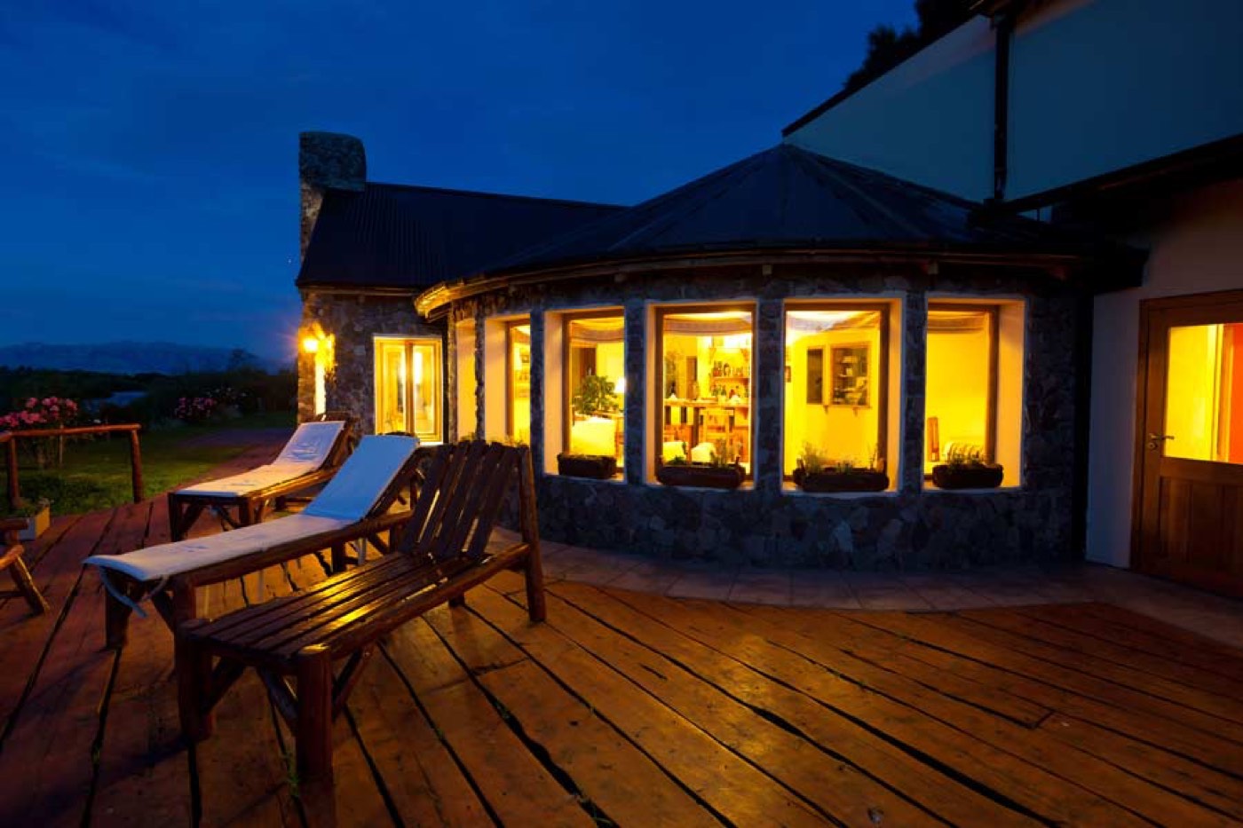 The exterior of a round stone-and-timber lodge wing lit up at dusk, lounge chairs set on the deck outside