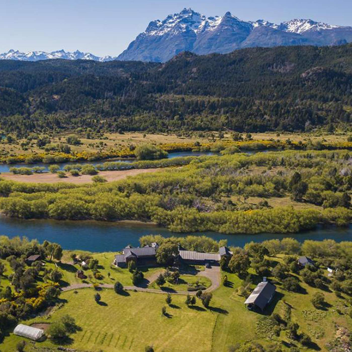 The lodge complex seen from above, beside a river bend framed by open pasture, poplar trees, and mountains under a clear sky