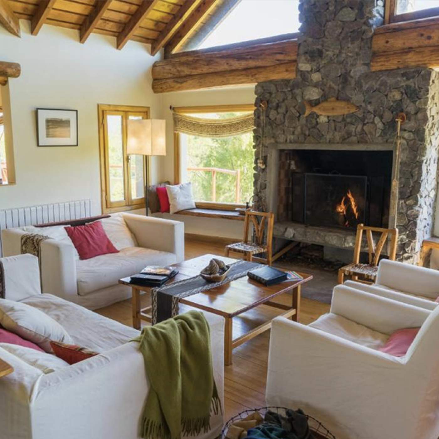 A bright great room with white sofas, a red accent pillow, a stone fireplace, and a window-seat bench overlooking the grounds