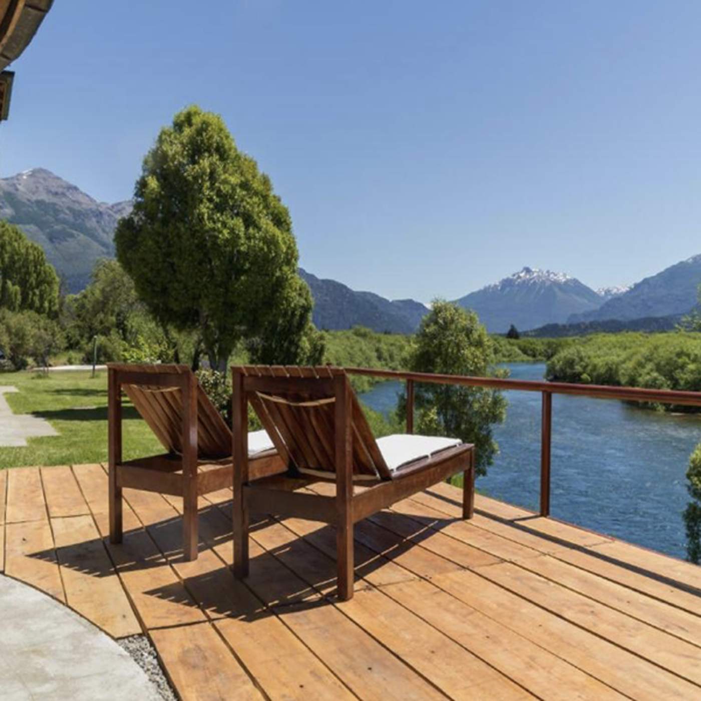 Two wooden lounge chairs on a deck overlooking a river, framed by trees, with a mountain range in the distance