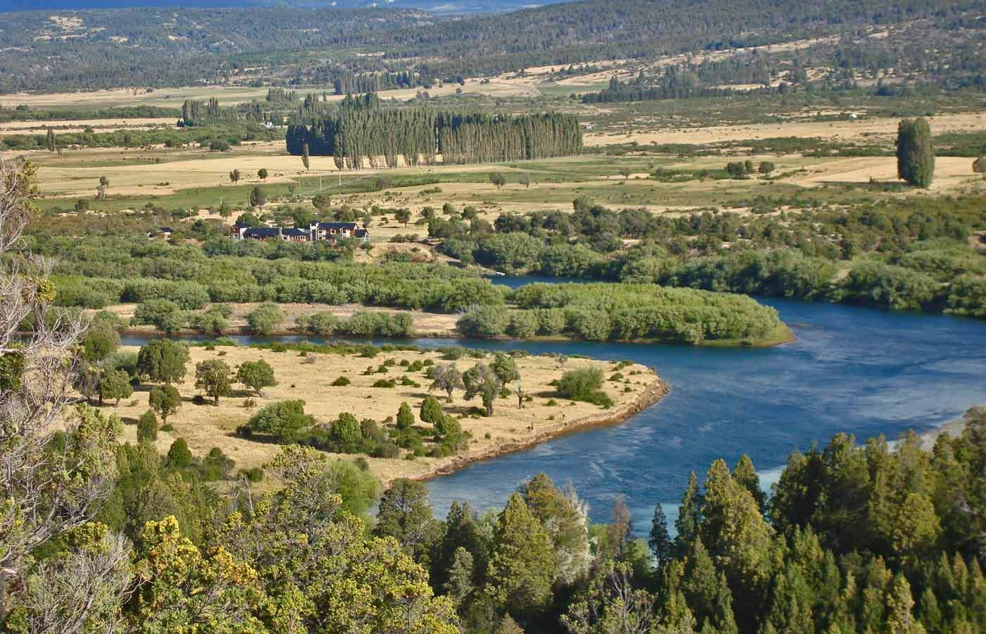 An elevated view of a river winding past farmland and a stand of poplars, the lodge visible in the distance against the mountains