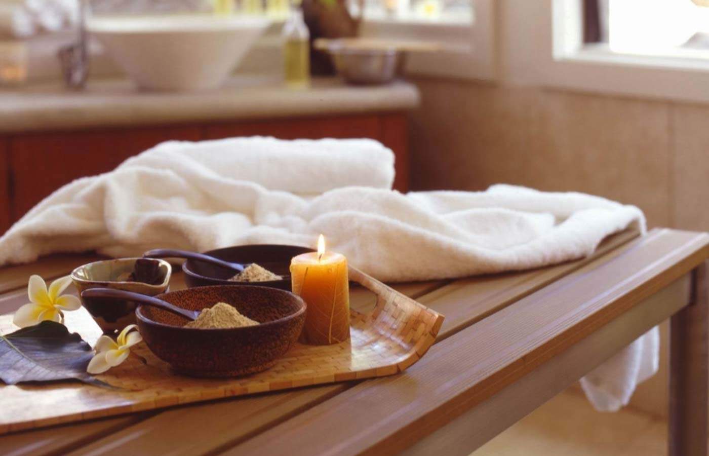 A spa tray of exfoliating scrubs, a lit candle, and frangipani flowers beside a rolled white robe