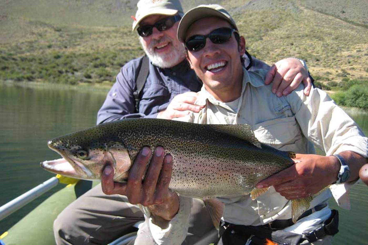 Two anglers on a boat together holding up a rainbow trout, hills rising behind them