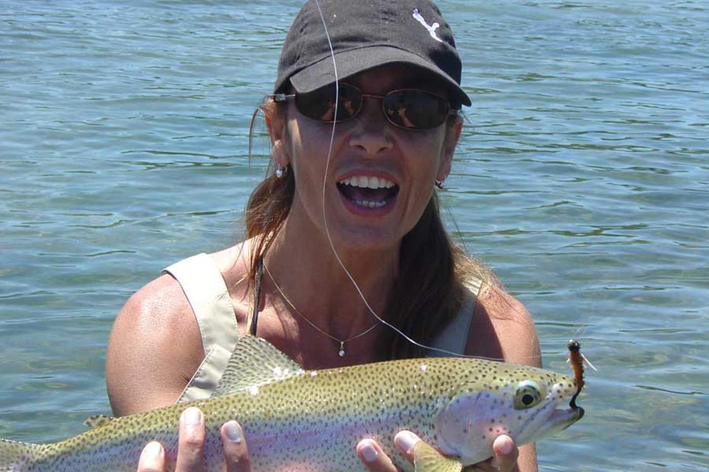 A laughing angler holding up a rainbow trout still hooked to the fly, standing in the shallows