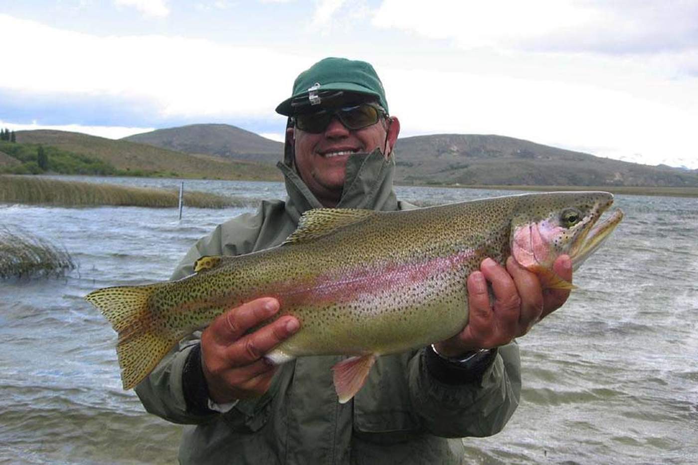 An angler in a green jacket holding up a large rainbow trout, rolling hills behind