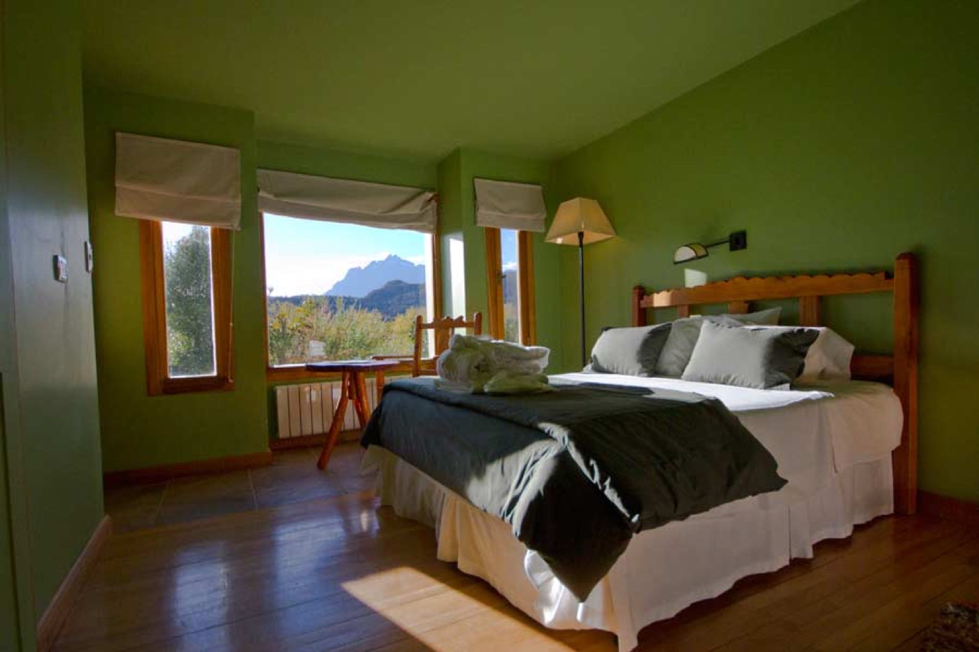 A green-walled bedroom with a wood bed frame, grey-and-white linens, and a mountain view through the windows